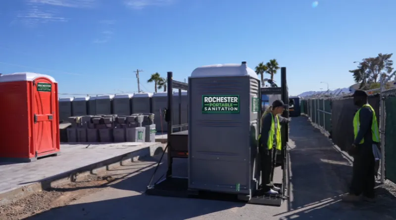 Two Rochester Portable Sanitation workers in safety vests setting a gray porta potty into place on a narrow paved lane in Rochester, NY